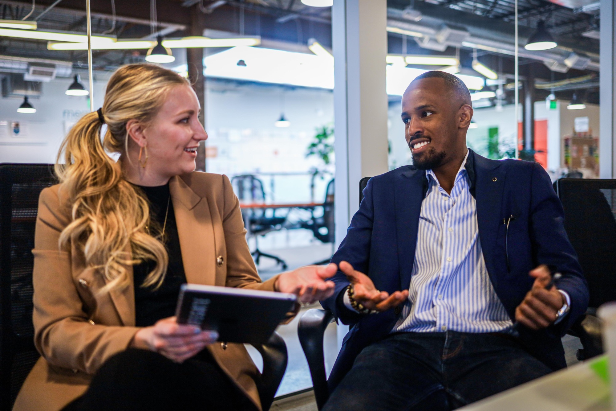 Two people - a white woman and a black man - meet in their office, both looking at an iPad and talking.